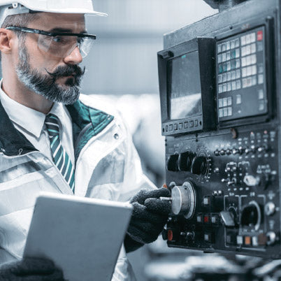 Engineer working on a machine with a tablet, wearing a white hard hat and safety glasses.